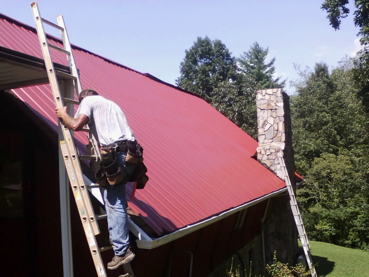 Roofer on a ladder beside a red metal roof and stone chimney — Soffit & Fascia Repair by Roofing Brothers in Coshocton